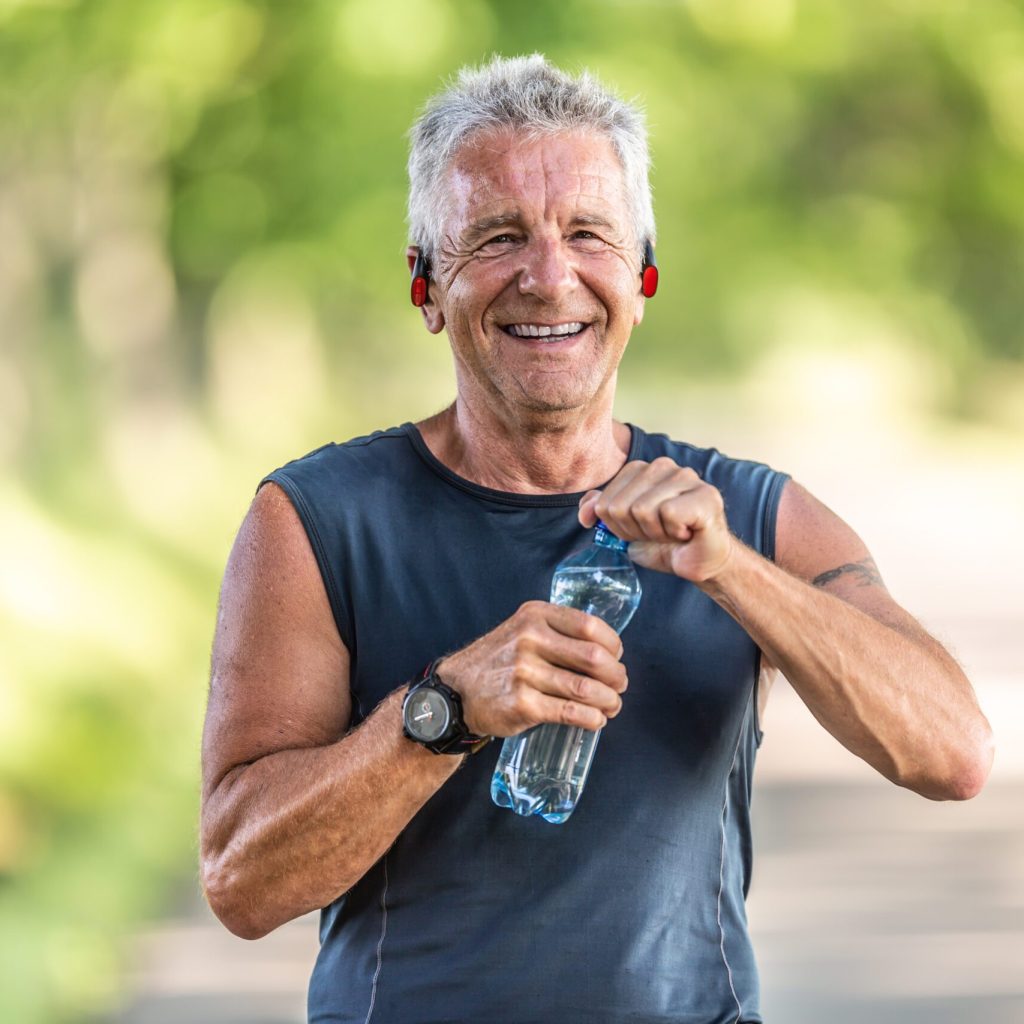 Smiling, fit and retired man with grey hair smiles as he opens up a bottle of water after finishing an aerobic workout.